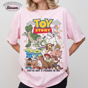 May include: A light pink short-sleeve t-shirt featuring a graphic print of Toy Story characters including Buzz Lightyear, Woody, Rex, and others. The text reads TOY STORY and YOU'VE GOT A FRIEND IN ME. The Comfort Colors Blossom logo is in the top left corner.