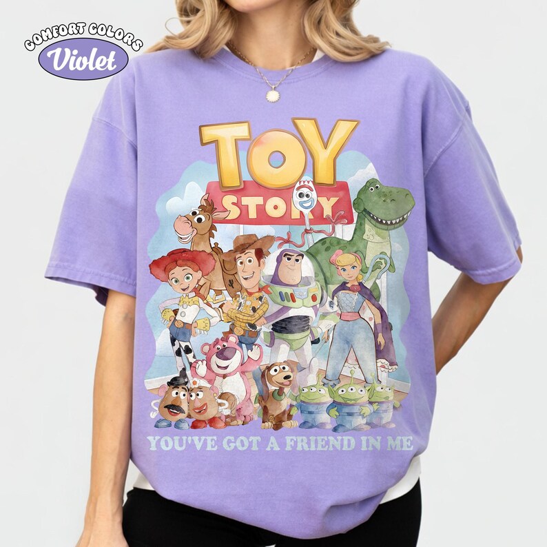 Puede incluir: Camiseta violeta Comfort Colors con un gr&aacute;fico colorido de personajes de Toy Story. El dise&ntilde;o incluye el texto "TOY STORY" y "YOU'VE GOT A FRIEND IN ME".