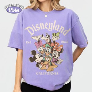 May include: A violet Comfort Colors t-shirt featuring a Disneyland graphic with various cartoon characters. The design includes the text "Disneyland," "Est 1955," and "California."