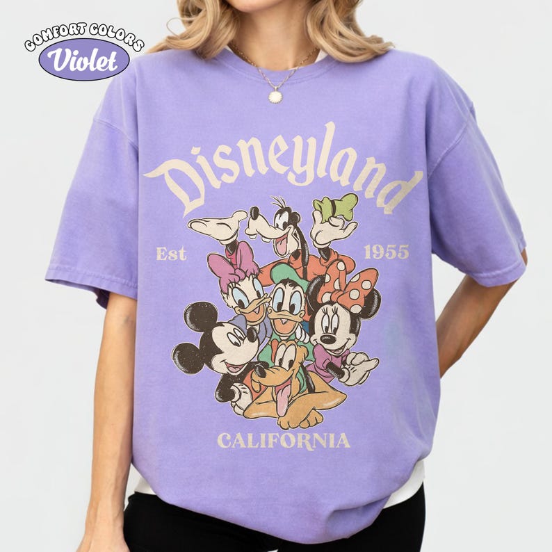 May include: A violet Comfort Colors t-shirt featuring a Disneyland graphic with various cartoon characters. The design includes the text "Disneyland," "Est 1955," and "California."