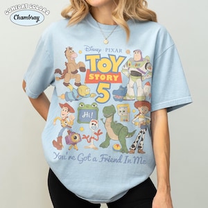 May include: Light blue t-shirt featuring a graphic of characters from Disney Pixar's Toy Story 5. The design includes Woody, Buzz Lightyear, and other characters. The text "You're Got a Friend In Me" is also visible.