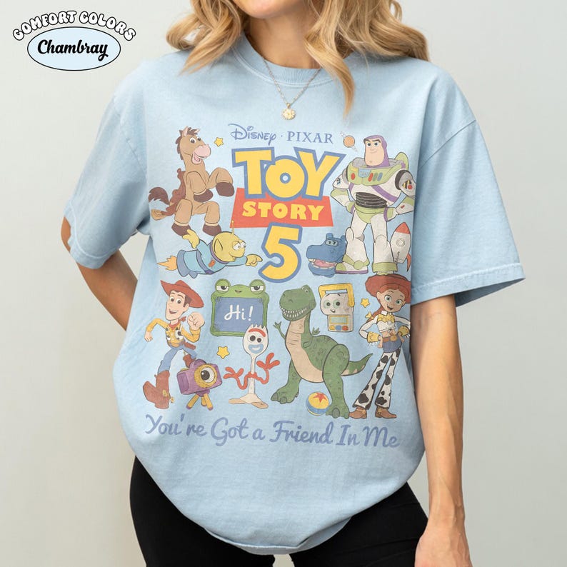 May include: Light blue t-shirt featuring a graphic of characters from Disney Pixar's Toy Story 5. The design includes Woody, Buzz Lightyear, and other characters. The text "You're Got a Friend In Me" is also visible.