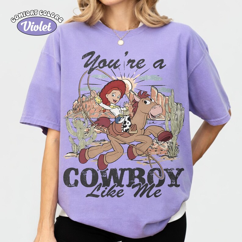 May include: A violet short-sleeve t-shirt featuring a graphic of a cowgirl riding a brown horse with a lasso. The design includes desert scenery with cacti, mountains, and the text "You're a COWBOY Like Me" in a distressed black font.