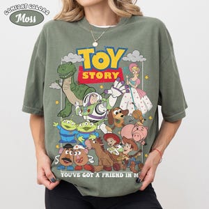 May include: A moss green short-sleeve t-shirt featuring a graphic print of Toy Story characters including Buzz Lightyear, Woody, Rex, and Bo Peep. The text reads TOY STORY and YOU'VE GOT A FRIEND IN ME. A Comfort Colors Moss label is in the top corner.