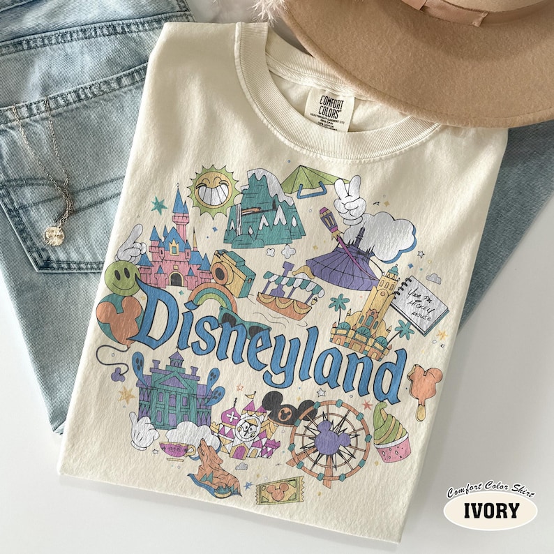 May include: Cream-coloured Comfort Colors t-shirt with a Disneyland graphic. The design features the castle, Matterhorn, and a Ferris wheel, with the word "Disneyland" in blue script. The shirt is ivory in colour.