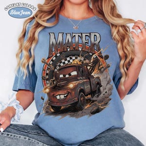 May include: Blue Comfort Colors t-shirt featuring a graphic of Mater from the movie Cars. The design includes the name "MATER" in a distressed font, a checkered flag, and lightning bolts. The shirt has the words "Comfort Colors" and "Blue Jean" printed on the top left.