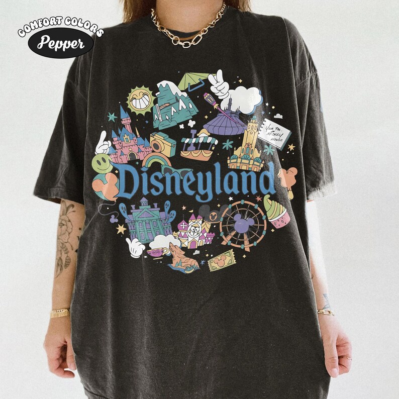 May include: Dark grey t-shirt featuring a colourful Disneyland graphic. The design includes iconic park elements such as a castle, Ferris wheel, and the Disneyland logo in blue. The shirt has a vintage, worn appearance, with the Comfort Colors and Pepper logos in the upper left corner.