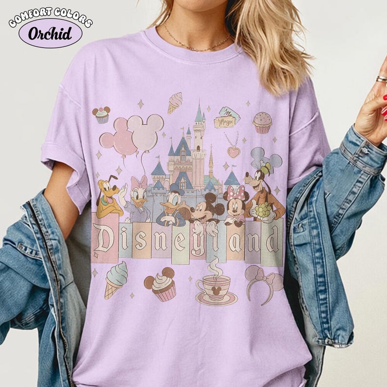May include: A light purple t-shirt with a Disneyland graphic. The design features cartoon characters, a castle, and the word "Disneyland" in pastel colors. The shirt also has the words "Comfort Colors" and "Orchid" in a circle.