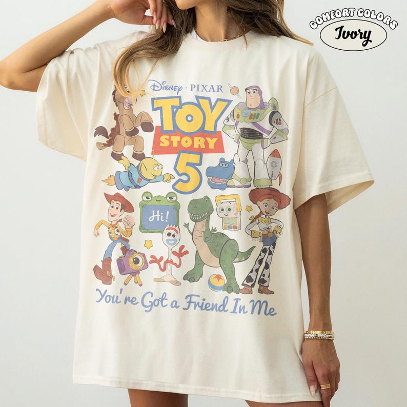 May include: Ivory-colored t-shirt featuring a graphic of characters from the Disney Pixar movie Toy Story 5. The design includes Buzz Lightyear, Woody, and other characters, with the text "You're Got a Friend In Me."