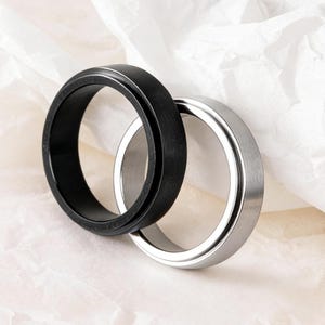 Puede incluir: Se muestran dos anillos, uno negro y otro plateado. El anillo negro tiene un acabado mate, mientras que el anillo plateado tiene un aspecto metálico brillante. Ambos anillos son circulares y parecen ser de metal.