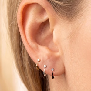 May include: Three small hoop earrings with clear, round, prong-set stones. The first hoop is gold-toned, the second is silver-toned, and the third is black. Each earring features a single sparkling stone centred on the front of the thin metal hoop.