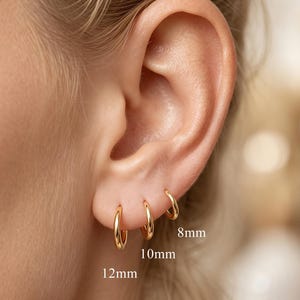 Könnte beinhalten: Drei goldene Creolen, die an einem Ohrläppchen getragen werden und mit den Größen 8mm, 10mm und 12mm beschriftet sind. Die Ohrringe sind dünn, kreisförmig und poliert und in einer Reihe von der kleinsten oben bis zur größten unten angeordnet.