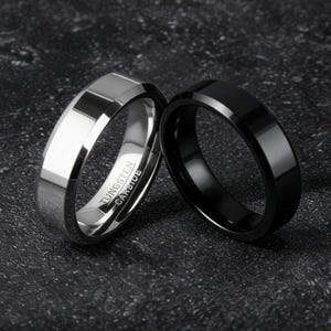 以下が含まれることがあります： 暗い質感のある表面に、銀色と黒色の2つの指輪が展示されています。 銀色の指輪はファセットデザインで、「Tungsten Carbide」の文字が入っています。 黒い指輪は滑らかで光沢のある仕上がりです。 結婚指輪またはファッションアクセサリーである可能性があります。
