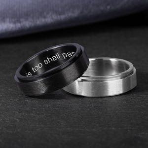 Op de afbeelding: Twee ringen, één zwart en één zilver, worden getoond. De zwarte ring heeft de inscriptie "is too shall pas" op de binnenband. De zilveren ring is effen. Beide ringen liggen op een donkere ondergrond.