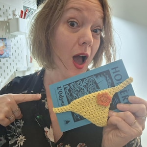 Può includere: Un perizoma giallo all'uncinetto con un fiore arancione è mostrato sopra un libro blu intitolato "L'Odyss". Il perizoma è realizzato con filato giallo chiaro. Il libro ha testo bianco e sfondo blu.