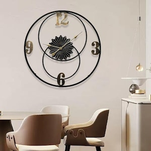 May include: A contemporary wall clock with a black metal frame and gold detailing. The clock has a circular design with overlapping rings, gold numerals at 3, 6, 9, and 12, and a decorative centre. The clock hands are gold.