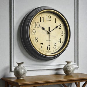 May include: A circular wall clock with a black frame and a gold inner rim. The clock face is cream-coloured with black numerals and hands. The word "QUARTZ" is printed on the face. The clock is mounted on a white wall.