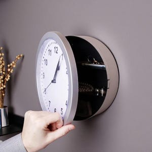 May include: A silver-framed wall clock with a white face and black hands, with a hidden compartment. The clock's door is ajar, revealing a dark interior. The clock is mounted on a grey wall.