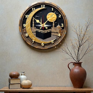 May include: A circular wall clock with a black face and gold Arabic numerals. The clock features a crescent moon, a sun, and a depiction of the Kaaba. The clock is framed in a brown wooden border. A vase with branches is on the right.