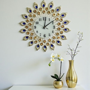 Puede incluir: Un reloj de pared decorativo con un marco de metal dorado, con detalles de joyas azules y naranjas. La esfera del reloj es blanca con números y manecillas negras. Un jarrón dorado y una pequeña maceta dorada con una planta están sobre una superficie blanca.