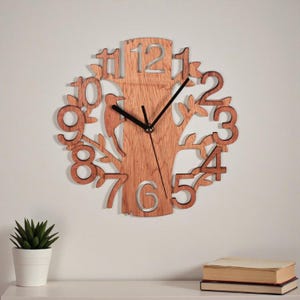 May include: Wooden wall clock with a tree design. The clock features a tree silhouette with cut-out numbers and a bird. The clock hands are black. The clock is mounted on a white wall.
