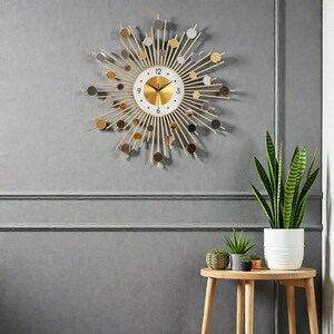 Può includere: Un orologio da parete dorato, stile raggiera, con quadrante bianco e numeri neri. Il design dell'orologio presenta linee radianti e accenti circolari in oro, argento e marrone. L'orologio è montato su una parete grigia.