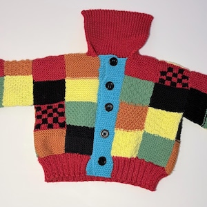 Könnte beinhalten: Ein farbenfroher Strickcardigan im Patchwork-Stil mit hohem Kragen. Der Pullover hat eine rote Basis mit Quadraten in Schwarz, Gelb, Grün, Orange und Blau. Er hat schwarze Knöpfe und rote Manschetten. Ein gemütliches, handgefertigtes Kleidungsstück.