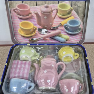 May include: A delightful ceramic tea set for children, including a pink teapot and cups adorned with white polka dots, saucers, and spoons. The set is presented in a decorative tin, with the text "Kids will go dotty for a cuppa from this charming ceramic tea set!"
