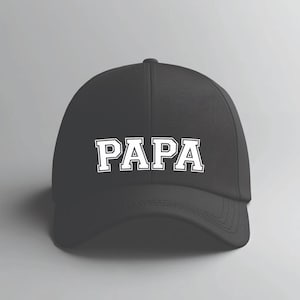 May include: Black baseball cap with the word "PAPA" in white, block letters across the front. The hat has a curved brim and a rounded crown. The text is outlined in a contrasting color.