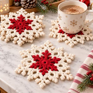 Könnte beinhalten: Ein Set aus drei gehäkelten Schneeflocken-Untersetzern mit cremefarbenem Außenrand und leuchtend roter Mitte. Ein Untersetzer trägt eine weiße Keramiktasse, die mit goldenen Schneeflockenmustern verziert ist, während die anderen flach liegen.