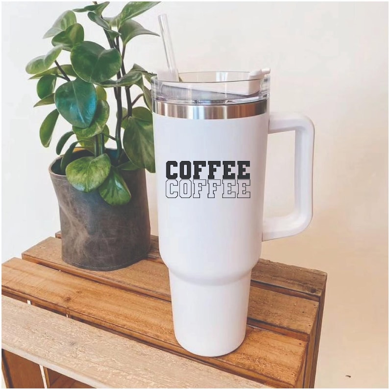 May include: White insulated coffee tumbler with a handle and the word "COFFEE" stacked twice in black. The tumbler has a clear lid and a straw. A small potted plant sits on a wooden crate in the background.