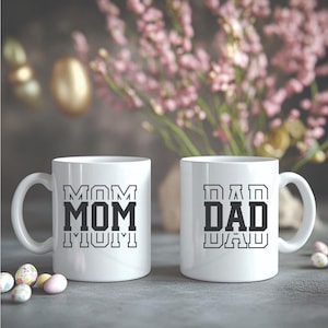 May include: Two white ceramic mugs with black text that reads "MOM" and "DAD". The "MOM" mug has the word repeated three times, and the "DAD" mug has the word repeated twice. The mugs are on a gray surface with Easter eggs and flowers in the background.