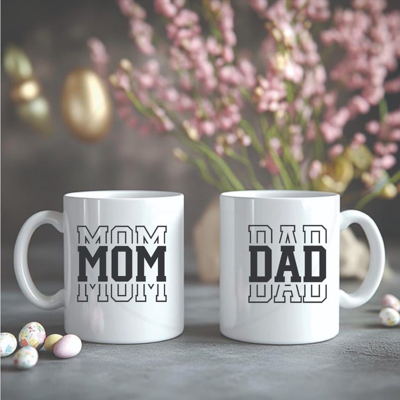 May include: Two white ceramic mugs with black text that reads "MOM" and "DAD". The "MOM" mug has the word repeated three times, and the "DAD" mug has the word repeated twice. The mugs are on a gray surface with Easter eggs and flowers in the background.