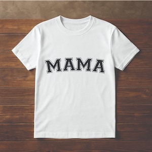 May include: White t-shirt with the word "MAMA" in large, black, block letters. The letters have a white outline. The shirt is displayed on a brown wooden surface.