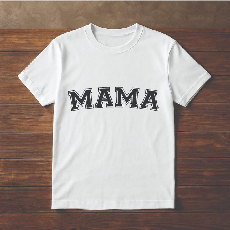 May include: White t-shirt with the word "MAMA" in large, black, block letters. The letters have a white outline. The shirt is displayed on a brown wooden surface.