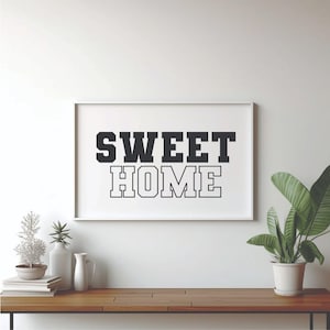 May include: A framed print with the words "SWEET HOME" in bold, black letters. The print is displayed on a wooden table, alongside decorative items and a potted plant with large green leaves. The background is a white wall.