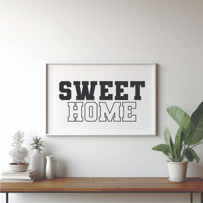 May include: A framed print with the words "SWEET HOME" in bold, black letters. The print is displayed on a wooden table, alongside decorative items and a potted plant with large green leaves. The background is a white wall.
