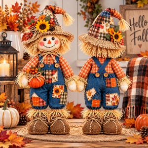 May include: Two scarecrow dolls wearing plaid shirts, denim overalls with fabric patches, and pointed hats decorated with sunflowers and berries. The dolls have straw hair, stitched facial features, and brown boots, standing on a round mat.