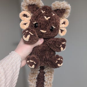 Könnte beinhalten: Ein handgefertigtes Axolotl-Plüschtier in Braun- und Beigetönen. Das Stofftier hat einen strukturierten, samtigen Körper mit helleren Akzenten an den Kiemen, Pfoten und am Schwanz. Es hat kleine, schwarze Knopfaugen und einen freundlichen Ausdruck.