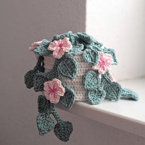 Könnte beinhalten: Ein handgehäkelter Blumentopf mit herabhängenden grünen Blättern und rosa Blüten. Der Topf ist weiß, und die Blätter sind ein gedämpftes Grün. Die Blüten haben rosa Blütenblätter und eine rosa Mitte. Der Topf steht auf einer weißen Oberfläche.