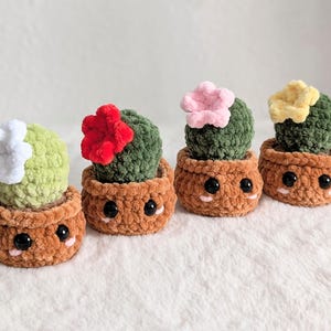 Könnte beinhalten: Vier handgefertigte Häkelkakteen in braunen Töpfen mit lächelnden Gesichtern. Jeder Kaktus hat eine andere Farbe, darunter Grün, und ist mit einer weißen, roten, rosa oder gelben Blüte gekrönt. Die Töpfe sind hellbraun.