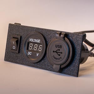 May include: A black rectangular mounting panel featuring a rocker switch, a circular digital voltage display labeled VOLTAGE DC V, and a circular USB charger port with a protective cover. Black electrical wires with red connectors extend from the back.