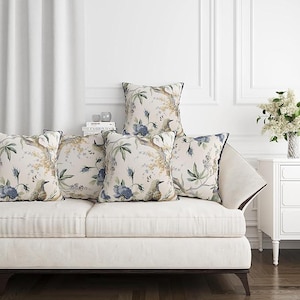 May include: Five decorative throw pillows on a white sofa. The pillows feature a floral and bird pattern in shades of blue, green, and beige, with a dark blue border. The sofa is in a bright, well-lit room.