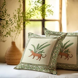 May include: Two square decorative throw pillows with a cream background. Each pillow features a detailed illustration of a leopard and green foliage. The pillows have a green and cream border with a repeating pattern.