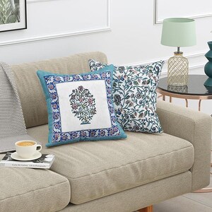 May include: Two decorative throw pillows on a beige sofa. One pillow features a floral design in a square frame with a teal border. The other pillow has a white background with a blue and green floral pattern.