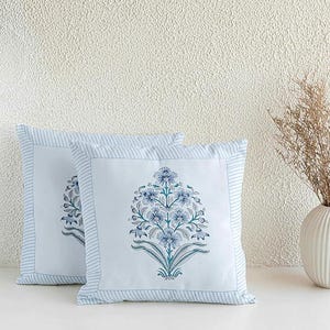 May include: Two square throw pillows with a floral design. The pillows have a white background with a blue floral pattern in the center. The edges are trimmed with a blue and white striped border. A vase with dried plants is in the background.