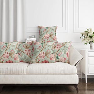 May include: Five decorative throw pillows with a floral pattern in shades of pink, red, and green on a light blue background. The pillows are arranged on a white sofa. The pillows have a pink border.