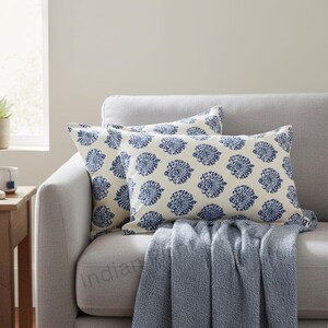 May include: Two rectangular throw pillows with a cream background and a repeating blue floral pattern rest on a gray sofa. A blue blanket is draped over the sofa. The pillows add a decorative touch to the living room.
