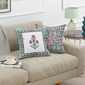 May include: Two decorative throw pillows on a beige sofa. One pillow features a white center with a pink floral design and a teal border. The other pillow has a teal background with a pink floral pattern.