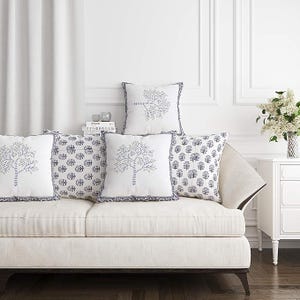 May include: A white sofa with five decorative throw pillows. The pillows feature blue tree and floral patterns. The pillows have a ruffled blue border. A vase of white flowers sits on a white side table.